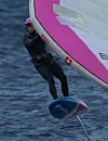 Seiler i våtdrakt på seilbrett med rosa og hvit seil på mørkt vann.