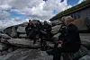 Fire personer sitter på store steiner ved kysten og snakker mens bølger og himmel danner bakgrunnen.