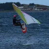Kitesurfer i våtdrakt og hjelm manøvrerer på urolig hav med kystbebyggelse i bakgrunnen.