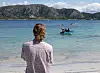 Person sett bakfra på strand med kajakker i en bukt og fjell i bakgrunnen.