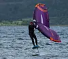 Vindsurfer i sort våtdrakt på et lilla foilbrett over bølger ved en skogkledd kyst.