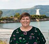 En smilende person står foran vann og fjell med en hvit bygning i bakgrunnen.