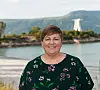 En smilende person står foran vann og fjell med en hvit bygning i bakgrunnen.