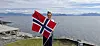 En person står ved vannet og holder et norsk flagg foran fjell og hav.