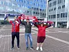 Tre personer står på en plass og holder opp supporter-skjerf foran en moderne bygning.