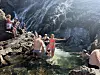 Flere mennesker bader og står i en liten kulp foran en mørk fjellvegg med foss.