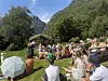 Folk sitter på gresset og på benker foran en liten scene i en fjellbygd.
