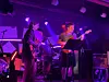 To musikere opptrer på en scene med lilla lys, mikrofonstativ og instrumenter.
