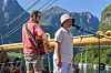 To musikere opptrer på en utendørsscene med mikrofon og gitar, med fjell og fjord i bakgrunnen.