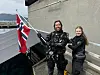 To personer står på et tak ved en grå vegg, mens den ene holder et norsk flagg.
