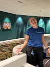 Smilende ansatt står ved kanten av en fisketank i et sjøtema-rom med blågrønne vegger.