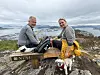 To personer sitter på en benk på et fjellplatå med utsikt mot sjø og by, med en liten hund foran.