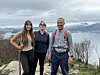 Tre personer står på en steinete utsikt med fjord og fjell i bakgrunnen.