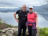 To personer står på en stein med ryggsekk og utsikt mot fjord, fjell og en by i bakgrunnen.