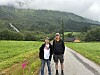 To personer står sammen på en grusvei med grønne marker og fjell i bakgrunnen.