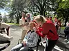 Folk i røde klær samles rundt en sittende person i en park med trær og sollys.
