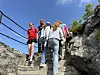 Flere personer går opp bratte steintrapper mellom store steiner og grønt i solskinn.