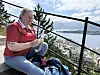Person i rød skjorte sitter ved et rekkverk med utsikt over by, fjord og fjell.
