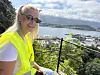 En kvinne i gul vest sitter ved rekkverket og ser utover en havn med fjell i bakgrunnen.