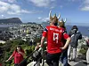 Flere personer står på en utsiktsplass med utsikt over en by, fjord og fjell under blå himmel.
