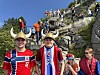 En stor folkemengde i røde klær står og sitter i en bratt steintrapp med fjell og blå himmel i bakgrunnen.