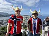 To personer i røde drakter og vikinghjelmer står foran et fjordlandskap.
