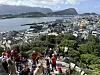 Stor folkemengde i røde klær står og går i en bratt trapp med by og fjord i bakgrunnen.