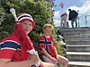 To barn i røde drakter sitter ved en trapp ute, med flere personer og et norsk flagg øverst i bildet.