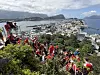 En stor gruppe mennesker i røde klær står og går langs en grønn fjellside med utsikt over en kystby og fjord.