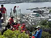 En gruppe mennesker i røde drakter sitter og står i en trapp med utsikt over havn og fjord.