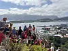 En folkemengde i røde drakter står på en ås med utsikt over en by og en fjord.