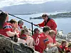 En gruppe mennesker i røde klær sitter på en steinrygg med utsikt over fjorden og fjellene.