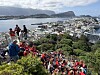 En stor folkemengde i røde drakter står og går i en bratt trapp med utsikt over en by og en fjord.