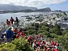 En stor folkemengde i røde drakter står og går i en bratt trapp med utsikt over en by og en fjord.