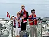 Tre personer står ved et glassrekkverk med et norsk flagg og utsikt over bygninger og vann.