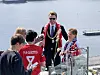 En gruppe mennesker i røde drakter og medaljer står samlet ved et utsiktspunkt med fjord i bakgrunnen.