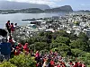 Stor folkemengde i røde drakter sitter og står i en bratt trapp med utsikt over Ålesund, havna og fjellene.