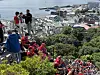 Stor folkemengde i røde klær sitter og står langs en bratt fjellside med utsikt mot havn og bygninger.