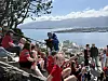En folkemengde i røde klær sitter og står på en skråning med utsikt over fjord og by.