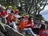En gruppe mennesker i røde klær sitter på steiner ved sjøen og ser på noe utenfor bildet.