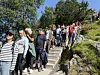 En rekke mennesker går ned en smal steintrapp langs en grønn bakke i solskinn.