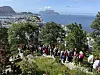 En stor folkemengde går langs en smal sti i grønt terreng med hav og øyer i bakgrunnen.