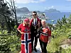 Tre personer står foran et utsiktspunkt med fjord, fjell og flere skip i bakgrunnen.