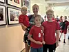 Tre barn og en voksen i røde trøyer står i en lys gang med bilder på veggene.