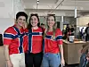 Tre kvinner smiler side om side i røde og blå supportertrøyer foran en kafédisk.