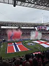 Et fullsatt stadion med store norske flagg på banen og røyk i rødt, hvitt og blått.