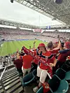 Supportere i røde klær står på tribunen med skjerf mens de ser ut over en fullsatt fotballstadion.