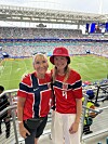 To kvinner i røde og blå norske fotballdrakter står på tribunen foran en fullsatt stadion.