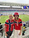 To kvinner i røde og blå norske fotballdrakter står på tribunen foran en fullsatt stadion.