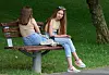 To ungdommer sitter på en parkbenk ved en gressplen og et tre.
